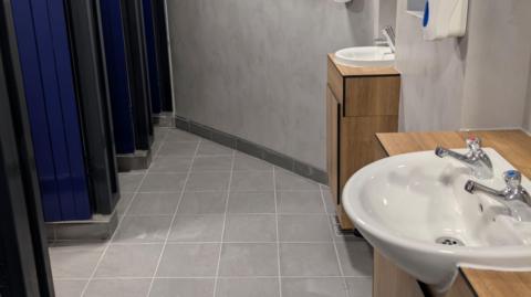 A clean, modern public restroom with a tiled grey floor and smooth light‑grey walls. Along the right side, there are two white sinks mounted on wooden cabinets, each fitted with chrome taps. A soap dispenser is mounted on the wall above the sinks. On the left side of the image, several cubicle doors in a dark blue colour are visible.
