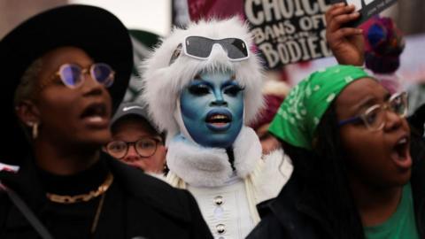 A demonstrator with blue face paint chants alongside two women amid placards at a protest.