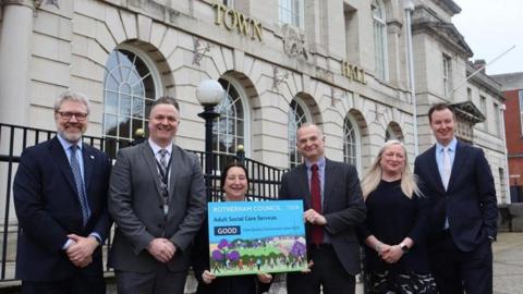 Four men and two women are standing in a line smiling at the camera. A woman and man n the middle are holding up a small posted which says "ROTHERHAM COUNCIL Adult social care services GOOD" They are standing in front of Rotherham Town Hall which is an off-white stone building with large arched windows.
