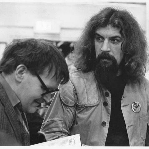 A black ad white snap of a bearded and long-haired Billy Connolly in 1981, looking at Alasdair Gray as he signs his book.