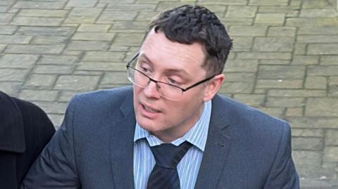 An image of a man with glasses wearing a grey suit and a blue and white striped shirt with a navy tie, walking on a concrete road.