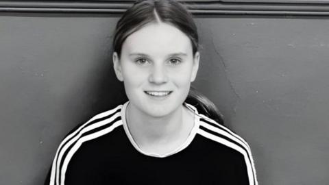 Black and white picture of Holly. She is sitting with her back against a wall and is smiling at the camera
