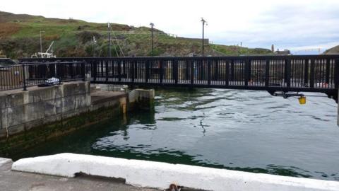 A black metal bridge with water underneath with a green hill in the background.