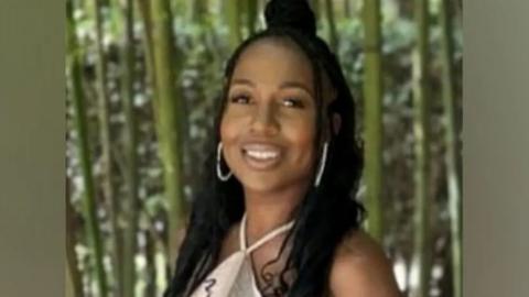 Michelle Sadio smiles at camera wearing large white hooped earrings and a pale coloured dress, in front of a green background.