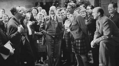 A black and white image showing Roger Livesey, in a kilt, and Michael Powell, seated, as the cèilidh scene is recorded in I Know Where I'm Going.