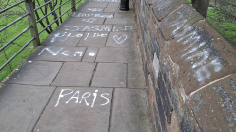 A stone walkway with a low wall on one side and a metal railing on the other. On the wall are words in white paint including 'Jasmine', 'Lilly' and 'Paris'. 