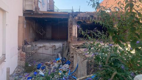 A disused building site with debris on the ground and overgrown weeds.