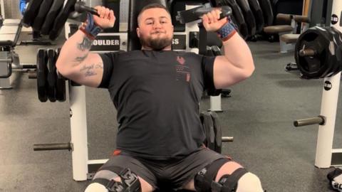 A man with dark hair and a beard in a black t-shirt and shorts is in a gym, lifting very large weights. There are large straps and supports around his legs