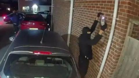 A photograph of a man wearing a face covering using some sort of electrical device near a car.