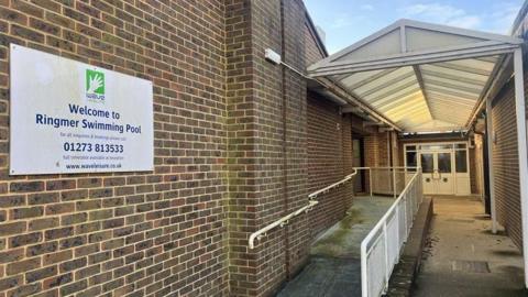 The entrance to a bricked-building. There is a white sign on the left which says "Welcome to Ringmer Swimming Pool".