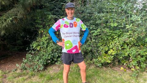 Kevin Webber standing outside against a bush wearing a black cap, Royal Marsden Cancer charity multi-coloured t-shirt and black shorts