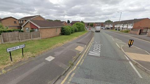 A picture of road  with houses on either side. There is also a pedestrian island with tactile paving and a sign with the road name. There is a white van and red car on the road in the distance. 