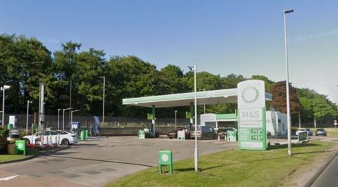A garage forecourt with vehicles parked at petrol pumps. The branding of the forecourt is green and white.