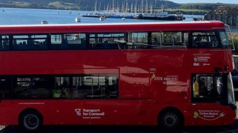 A red double decker electric bus from Go Cornwall Bus. It is parked on a road overlooking a river which has several yachts on it. A driver is sat at the front of the bus. A couple of passengers are sat inside the bus.