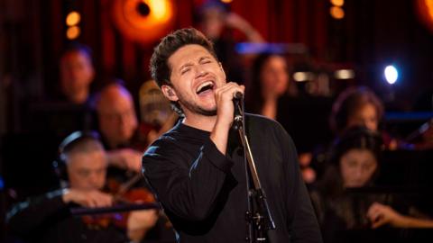 Niall Horan wearing a black top, singing in front of the BBC Concert Orchestra