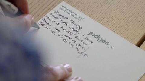 An image of someone writing a postcard from Judges Postcards in Hastings, East Sussex. The postcard has the Judges logo and says 'when did you last send a postcard?'.