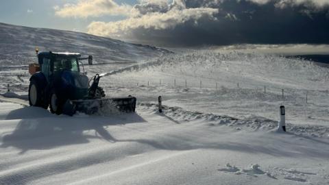 A tractor-mounted snow blower clearing part of the carriageway on the mountain. The landscape around it is white with snow.