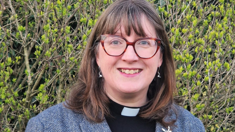 A head and shoulders shot of Kate Massey who is wearing brown glasses and a grey jacket. Behind her is a hedge of brown branches with green leaves. She has shoulder-length brown hair and smiles at the camera.