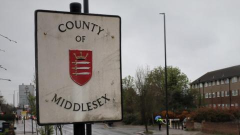 A white street sign reading COUNTY OF MIDDLESEX with a red coat of arms in the centre. The sign is beside a main road