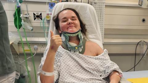 Rebecca O'Malley in hospital wearing a medical gown and a neck brace. She gives the camera a thumbs up and a faint smile. 