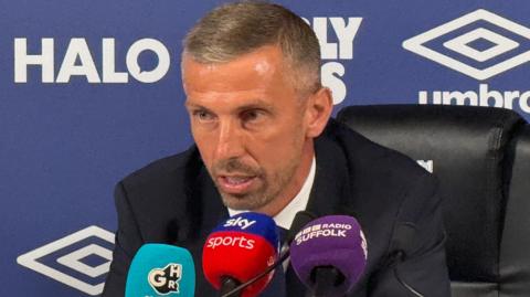A man wearing a suit is sat in front of three microphones with a blue sports press conference background behind him