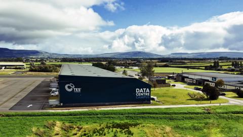 A aerial view of a warehouse type building that is navy. It says "data centre" in white writing along the side. Behind it are rolling hills set against a sky with some blue and some clouds. the building is in a semi-rural area surrounded by green fields with one other smaller white industrial building to the right.