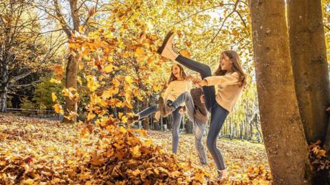 Three children kick colourful yellow and orange leaves outdoors in a park
