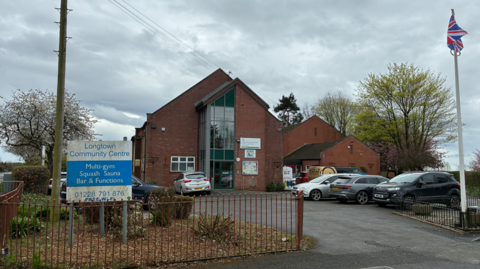 An exterior view of Longtown Community Centre. The building is a large, red brick structure with a long vertical window, iron railings and a sign outside with the centre name on it. 