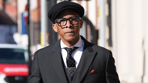 Image shows Jay Blades arriving at Worcester Crown Court wearing a black suit, white shirt, black tie with white spots and a tie pin and a red handkerchief tucked into his breast pocket. He is wearing a black peaked cap.