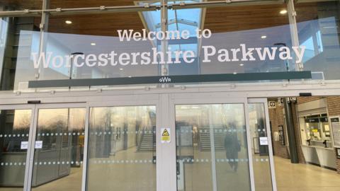 The entrance to Worcestershire Parkway station with a sign above the glass doors, a ticket office to one side and a passenger walking through the foyer.