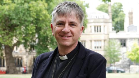 A man with short grey hair and smiling is wearing a clerical white collar with a black shirt and black jacket. He is standing in front of a large stone building and a tree that have been blurred out.