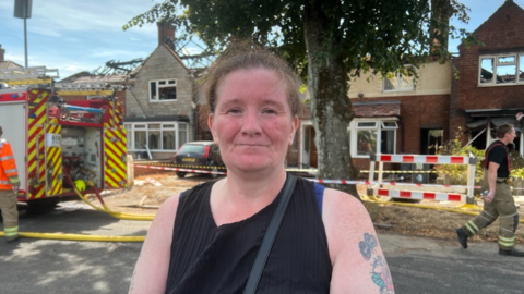 A woman with brown hair tied back with tattoos on her left arm. She is wearing a black tank top and is standing in front of a fire engine which is outside a house damaged by fire with no roof