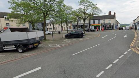 A Google Street view image from a junction showing a car park on the left with trees and two vehicles parked up. A road curves round to the left behind the car park. High street shops line the side of the street.