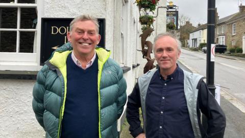 Two men with grey hair standing in front of a Pub in Grampound - One wears a green jacket and the other a black jacket