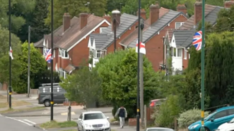 Flags hang from lampposts in a Birmingham street in between houses. The road is steep with cars parked on drives and in the road. A person walks up the hill.