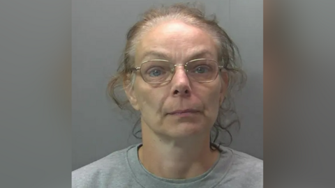 Custody photo of Melanie Wright. She has curly greying hair that has been tied back from her face and is wearing thin framed gold rimmed glasses. She is also wearing a plain grey sweatshirt.