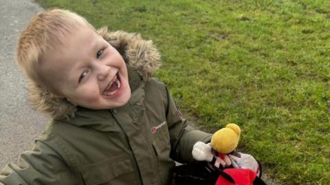 William, nine, has blond hair and is wearing a dark green Berghaus parka. He is holding toys including Mickey Mouse and Bullseye from Toy Story. He has a huge grin on his face and is standing on a path near some grass. 