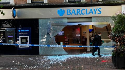 A Barclay's storefront. A large glass window has been smashed and red paint is visible on the pavement in front. Police tape has blocked off access to the bank. An officer in gloves and a mask is leaving the store.