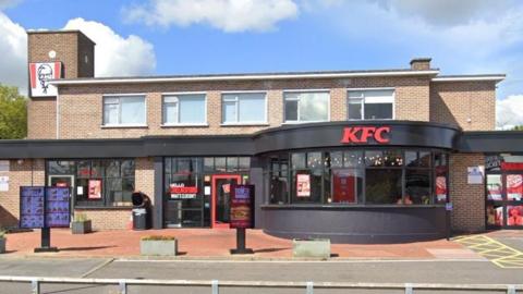 The exterior of a KFC restaurant in Broomfield Road, Chelmsford. It has a black trim frontage. The KFC branding is on the ground floor, but there is also an upper storey. There is blue sky and clouds above.