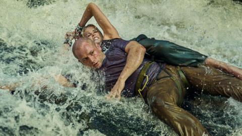 Egerton and Theron both cling to a large bolder in a river, with water crashing over them. Egerton wears a blue t-shirt and brown trousers, while Theron wears green trousers.