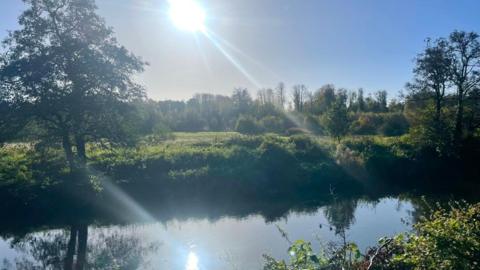 The sun shines brightly in the blue sky and is reflected in the river below with trees and fields 