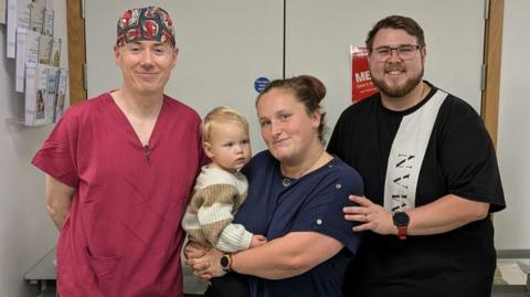 Consultant of paediatric urology Mark Woodward, with patient Parker and his parents Sophie and Matthew. Mr Woodward is wearing red hospital scrubs. Sophie is stood next to him, holding Parker, while Matthew stands next to Sophie with his hand on her shoulder. Everyone except Parker is looking and smiling at the camera. They are pictured inside a hospital. 