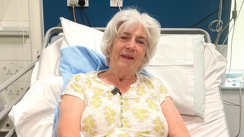 A woman smiling at camera lying in a hospital bed. White and yellow blouse, grey hair and wearing a hospital name wristband.