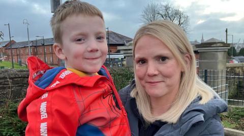 Terri Lee-Watson is lifting up her son Jace. She has blonde hair and is wearing a grey padded coat. He has short brown hair and is wearing a Spiderman jacket. The backdrop is a red brick building.