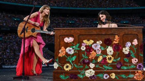 Taylor Swift and Gracie Abrams perform on stage during "Taylor Swift | The Eras Tour" at Wembley Stadium on June 23, 2024 in London. Taylor wears a flowing bright pink dress and holds an acoustic guitar, kicking out her right leg. Gracie sits behind a piano painted with flowers, she has short brown hair and brown eyes. 