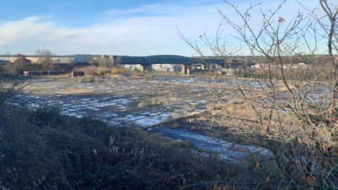 The former glassworks site at Deptford Terrace in Sunderland. The site is empty, with the ground covered in concrete paving. Grass and plants have grown across the land. The Queen Alexandra Bridge can be seen in the background.