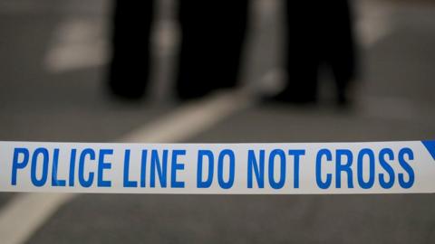A close up of police tape that reads "police line do not cross".  The blurred legs of two people are in the background.