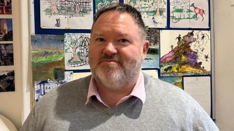 A man with short brown and grey hair, with grey facial hair. He is standing in front of a board with colourful paintings and drawings on it. He is wearing a grey jumper with a pink shirt underneath.