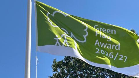 A green flag with a white stripe across the bottom and a tree with a parent and child stood next to it in white. It reads 'Green Flag Award 2026/27. A tree is in the background against the blue sky.