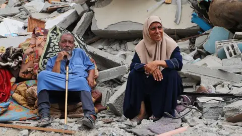 An elderly couple grieves in front of their destroyed home as Palestinians, carrying what belongings they could with them, returning to the Shuja'iyya neighborhood after the ceasefire agreement came into effect are seen walking among buildings destroyed in attacks by the Israeli army on October 16, 2025 in Gaza City, Gaza.